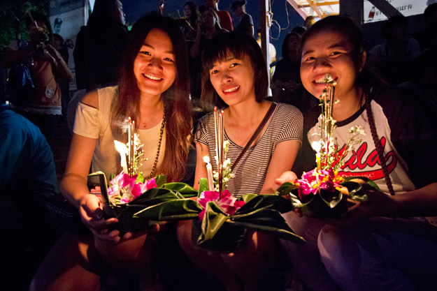 Three Thai girls at the riverside prepare to launch their Loi Krathong