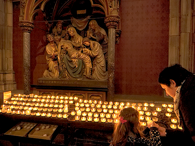 Mother helps her daughter light a candle at Cologne Cathedral. Behind the candles is the 13th Station of The Cross, a sculpture by Wilhelm Mengelberg illustrating Christ's body being taken down from the cross.