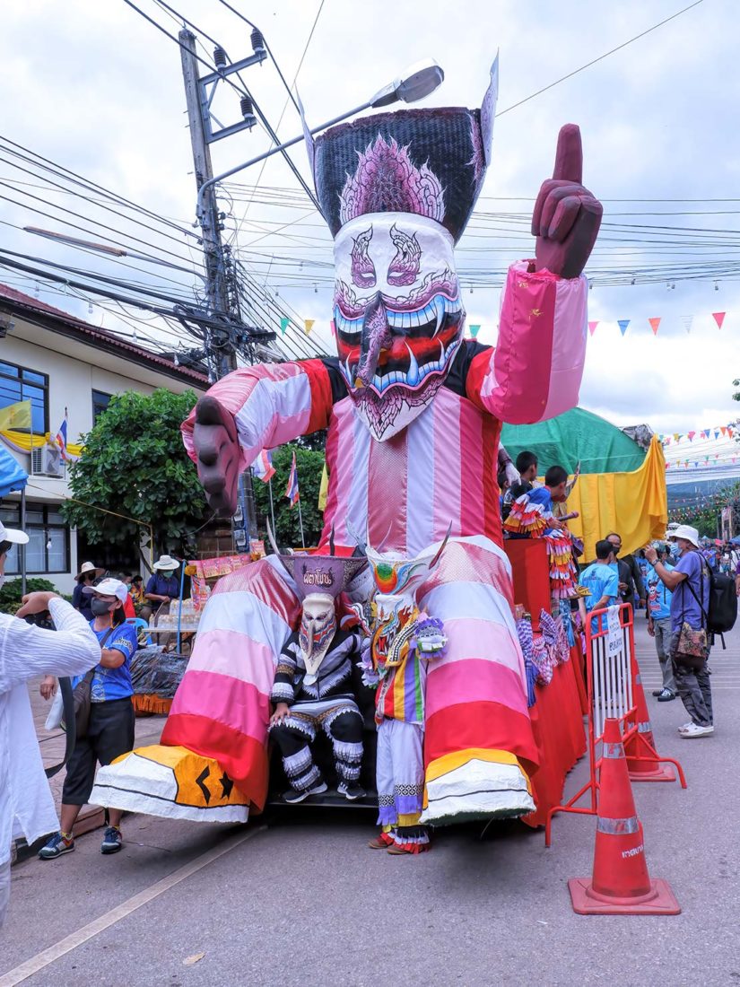 Costumed man poses in front of a parade float that features a Phi ta Khon Yai