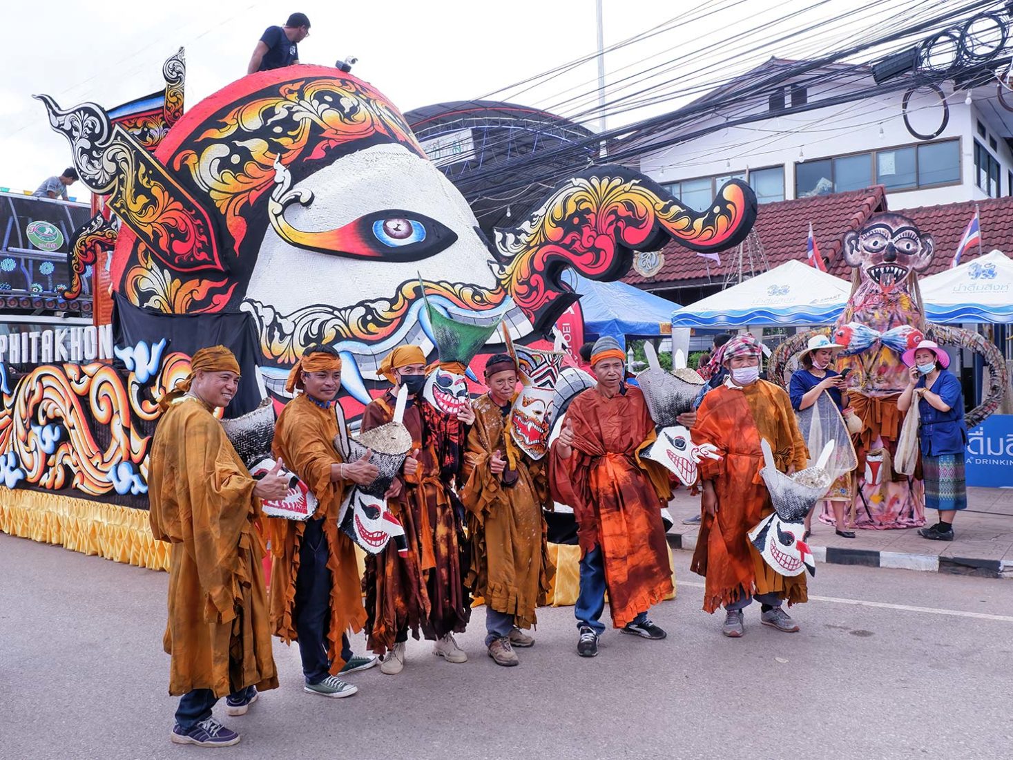 Local men in costume pose in front of a parade float that features a huge Phi ta Khon mask